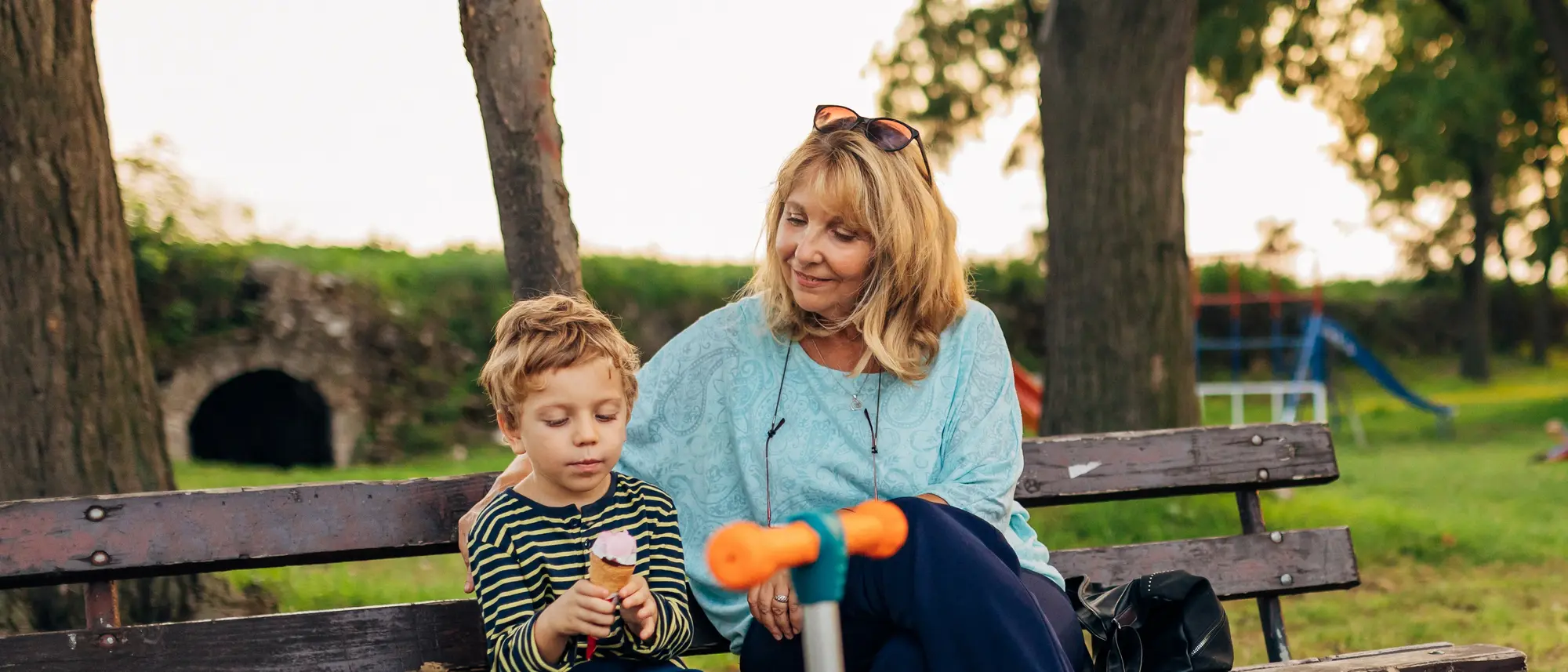 Eine Frau und ein kleiner Junge sitzen zusammen auf einer Bank im Park zu sehen. Der Junge hält ein kleines Spielzeug, während die Frau ihn freundlich anschaut. Vor der Bank steht ein orange-blauer Roller.