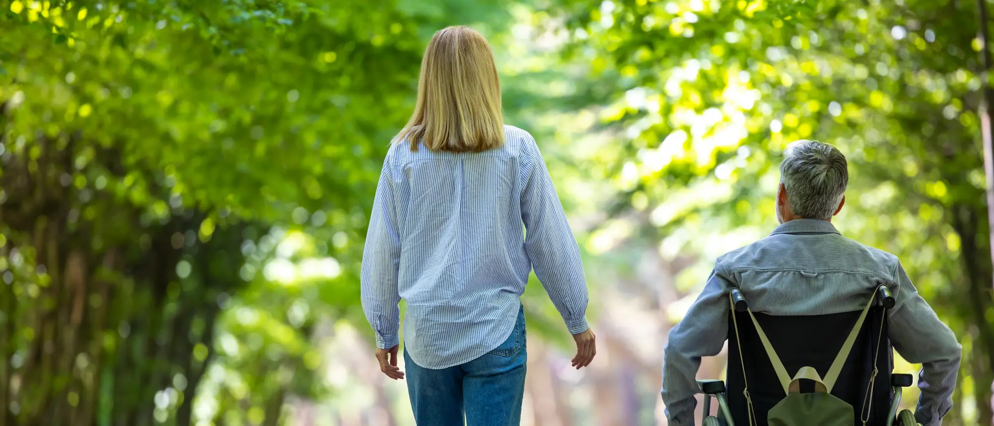 Eine Frau läuft neben einem Mann im Rollstuhl. Sie gehen einen breiten Weg entlang, der von Bäumen auf beiden Seiten umgeben ist. Der Hintergrund zeigt grüne Blätter, durch die das Licht der Sonne scheint.